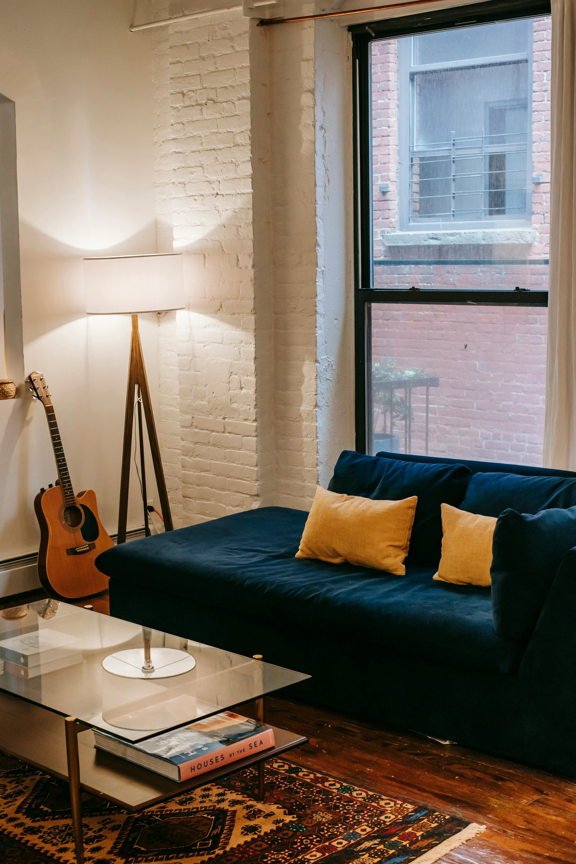 Un sofá cómodo con cojines amarillos, situado cerca de una ventana y una mesa de cristal, en una habitación acogedora con una lámpara y una guitarra, dentro de un apartamento.